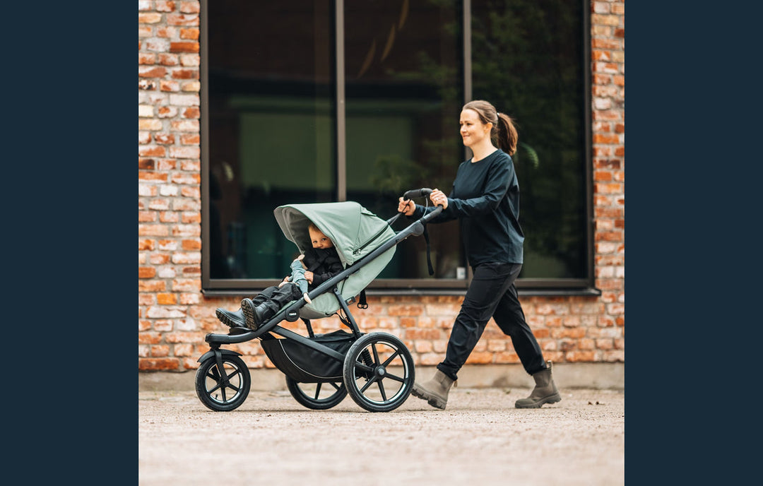 Caregiver walking with a child in a Thule Urban Glide 3 stroller beside a city brick wall.