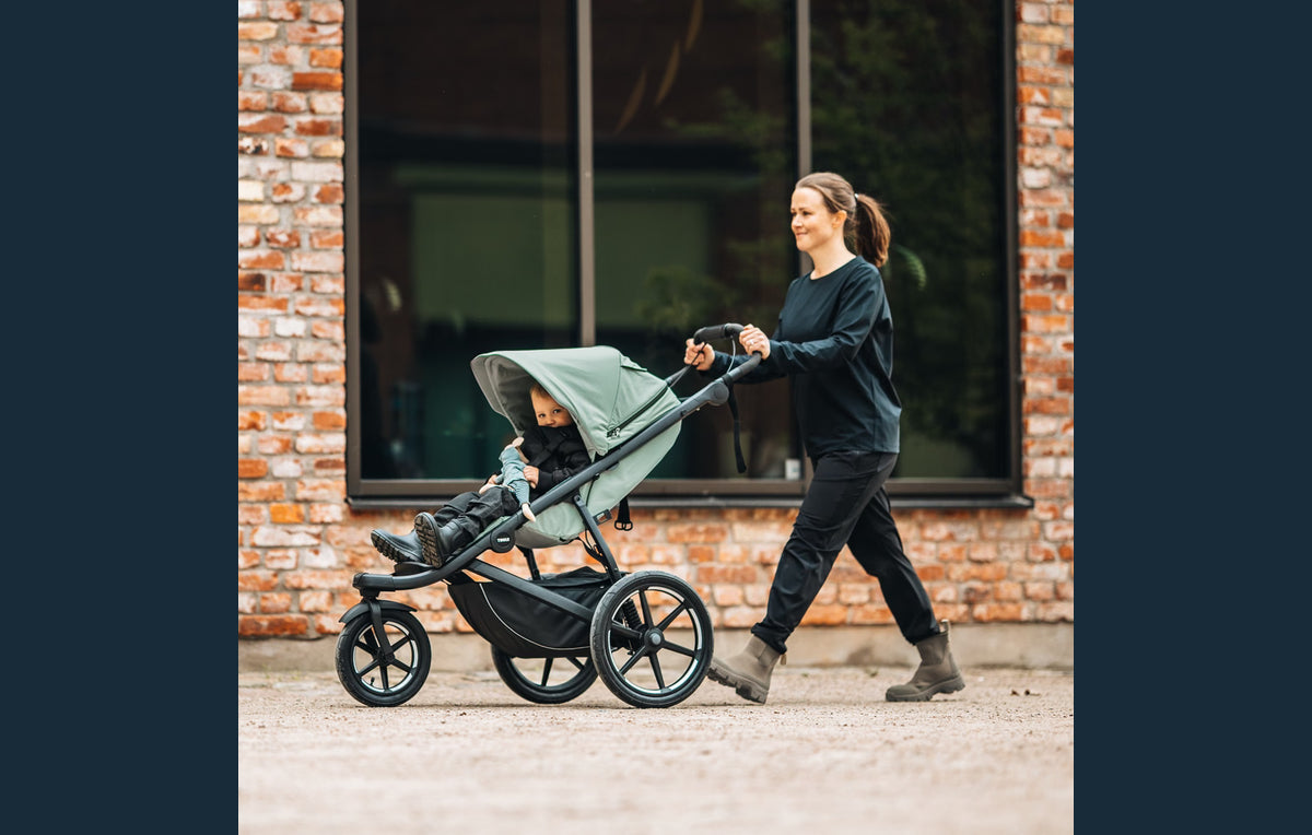 Caregiver walking with a child in a Thule Urban Glide 3 stroller beside a city brick wall.