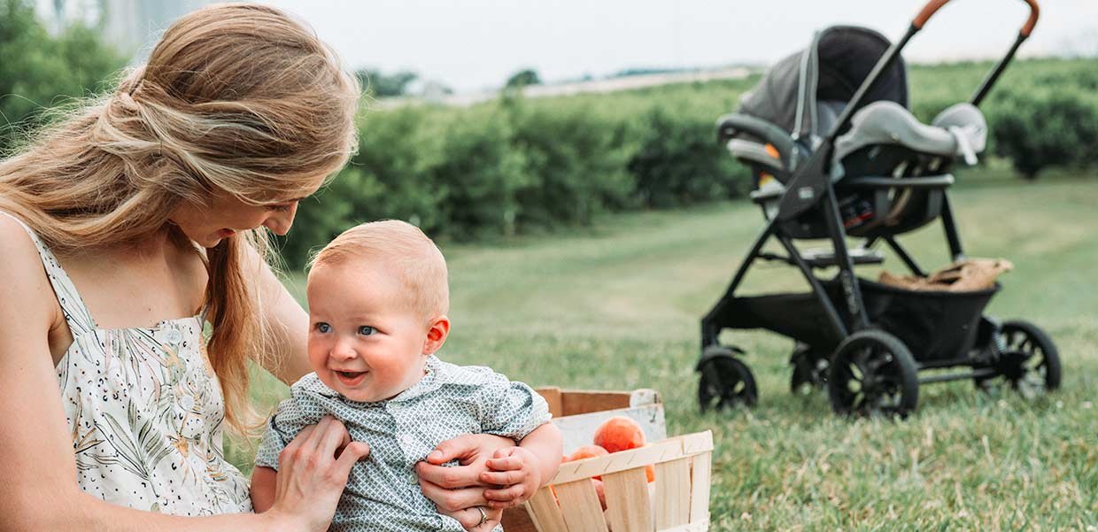Mother holding her baby in an orchard with a Chicco travel system