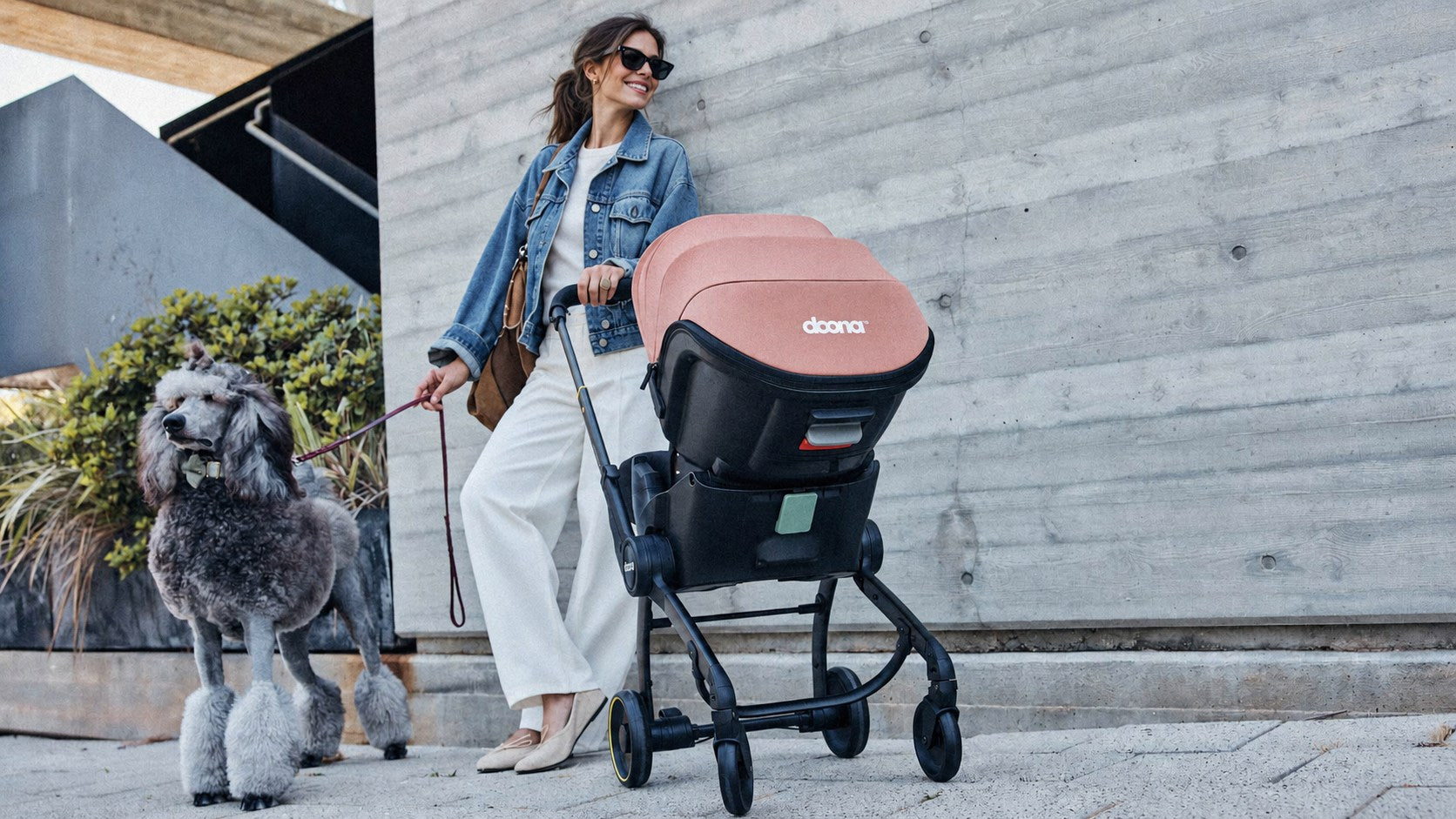 A young mother walking her dog with a Doona X stroller in Terracotta beside a concrete city building
